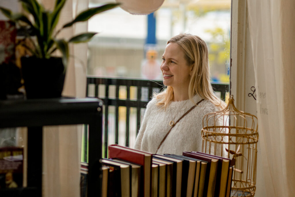 Adelaide Clemens smiles through a window standing near a book store in a scene from The Swearing Jar