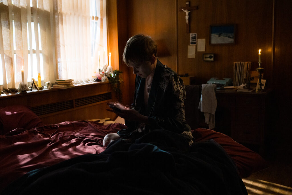 The narrator of I hate people, people hate me, sits on the bed of a dark room in a scene from I Hate People, People Hate Me