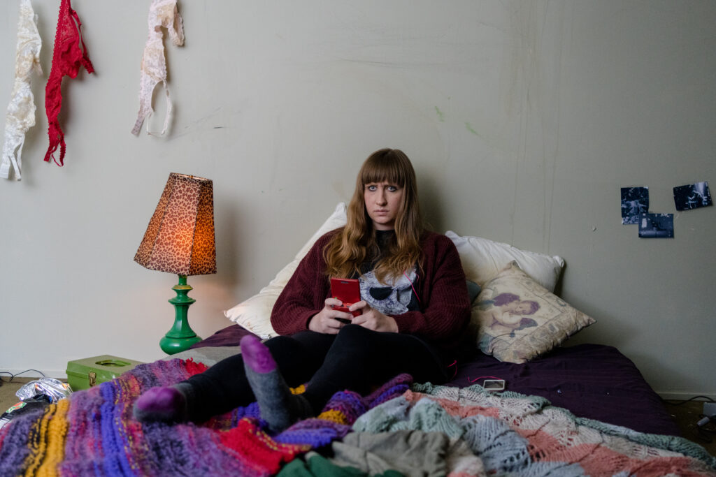 Lily Kazimiera sits in her bed in her room and looks at her phone in a scene from I Hate People, People Hate Me