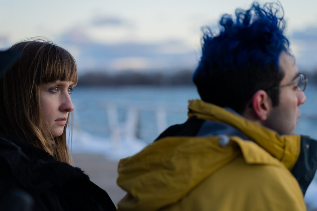 Lily Kazimiera and Bobby Summer sit on a bench in the winter along a lake and talk in a scene from I Hate People, People Hate Me