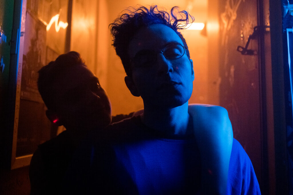 Bobby Summer looks to camera with blue light washing over his face in a scene from I Hate People, People Hate Me