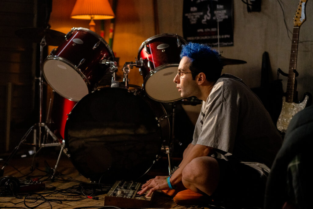 Bobby Summers sits on the floor of his bedroom playing keyboard with the drums in the back in a scene from I Hate People, People Hate Me