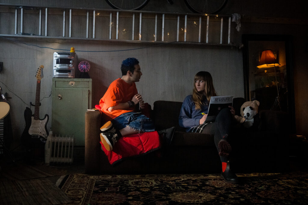 Bobby Summers and Lily Kazimiera sit on a coach and talk to eachother in a scene from I Hate People, People Hate Me