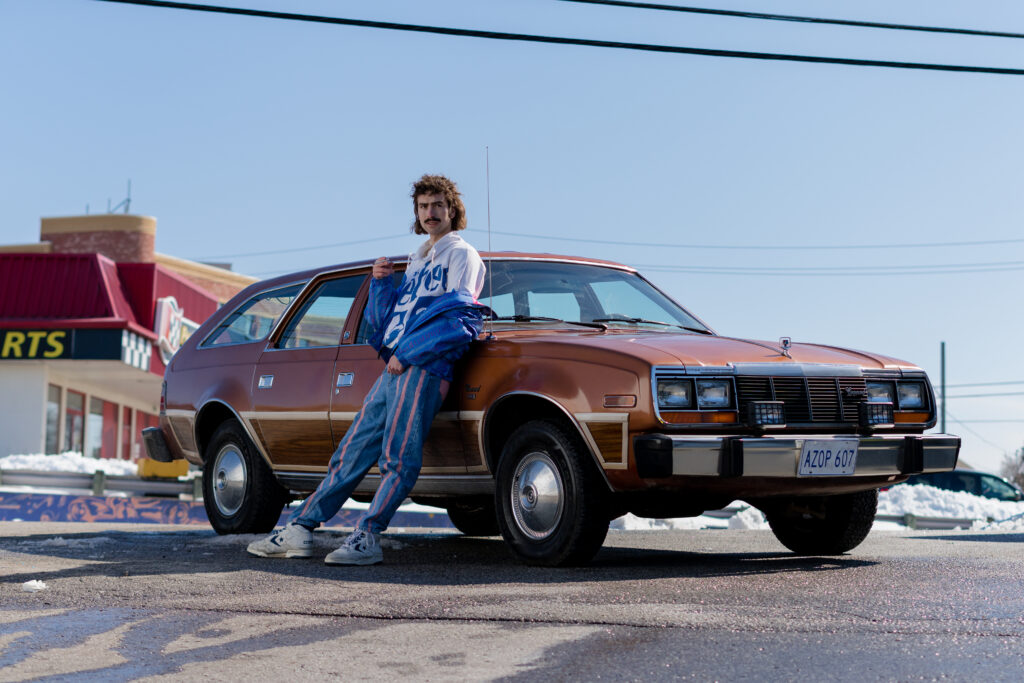 Lily Kazimiera love interest stands by a brown car in a scene from I Hate People, People Hate Me