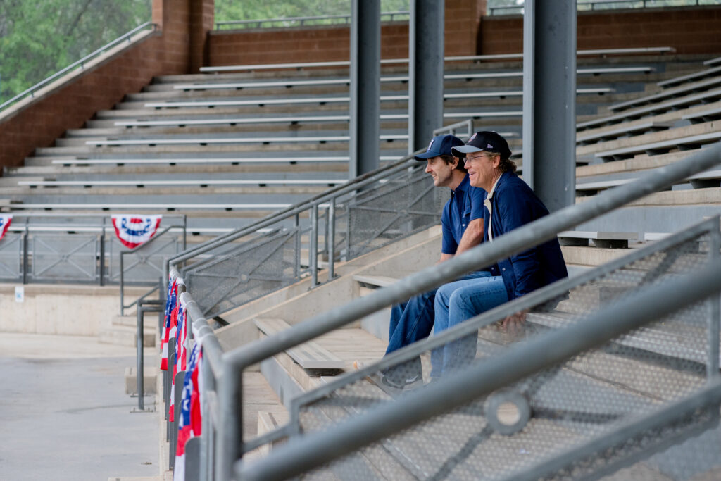 Greg Kinnear and Luke Wilson talk seriously together in baseball stadium