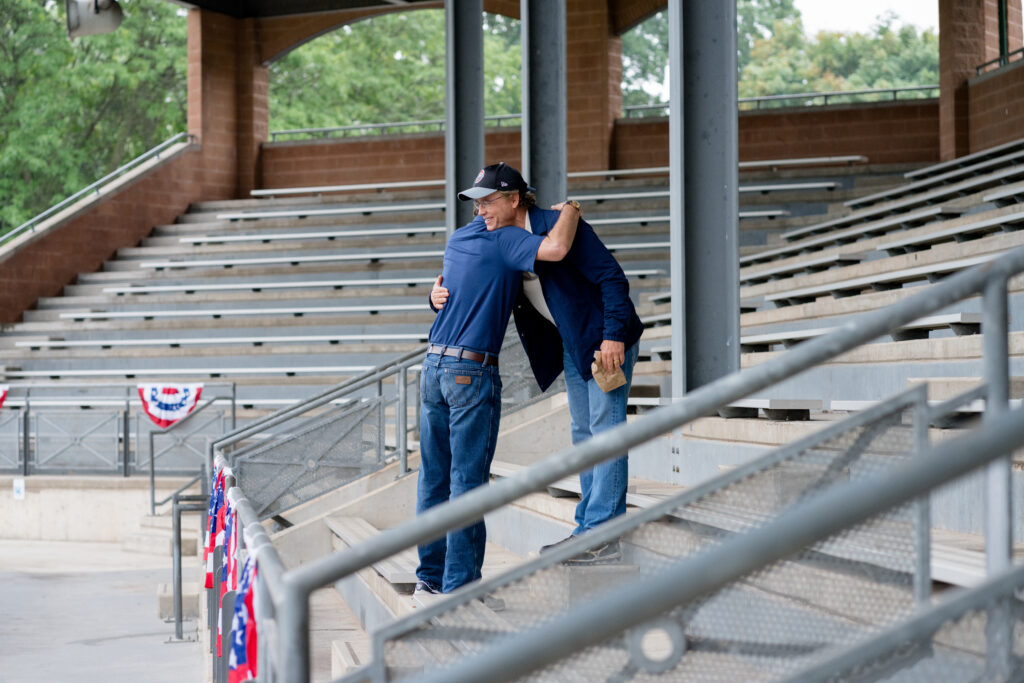 Greg Kinnear and Luke Wilson embrace as they say hello in baseball stadium