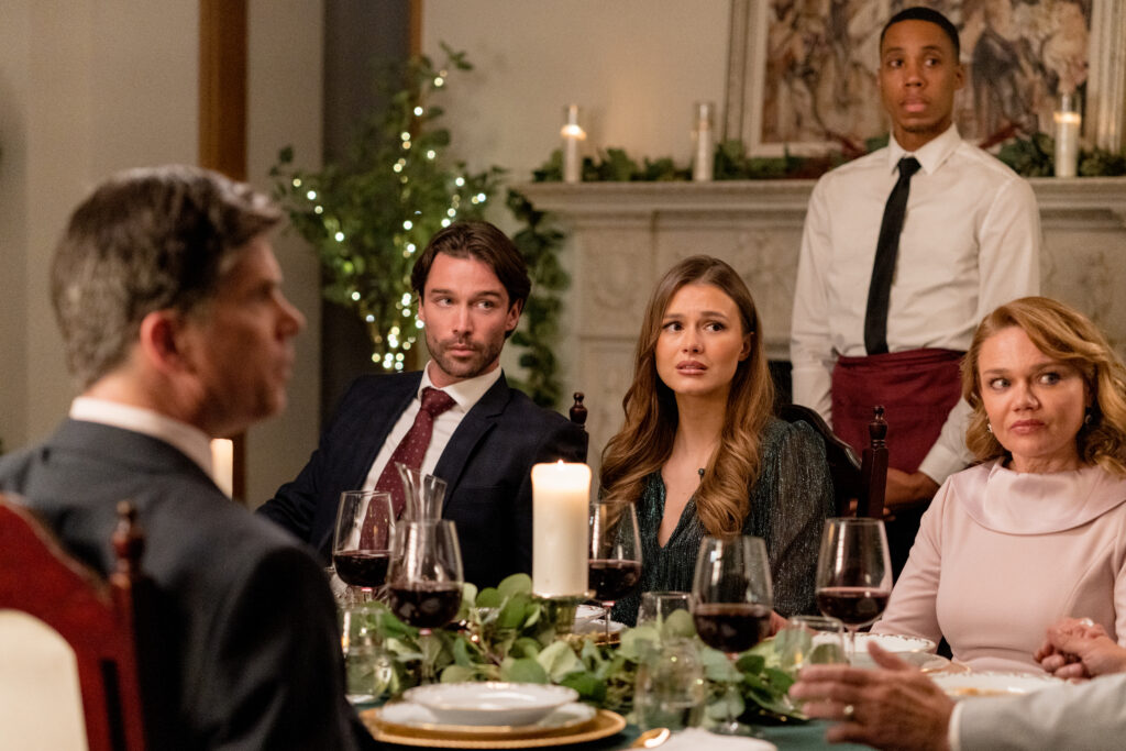 Celeste Desjardins and Joe Scarpellino sit at a dinner table looking worried in a scene from the Hallmark movie Christmas with the Knightlys