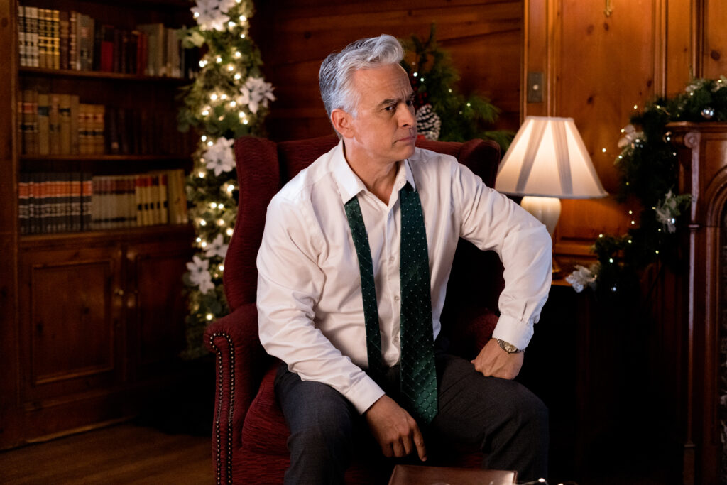 Randy Thomas sits in a library decorated for christmas in a white shirt in a scene from the Hallmark movie Christmas with the Knightlys