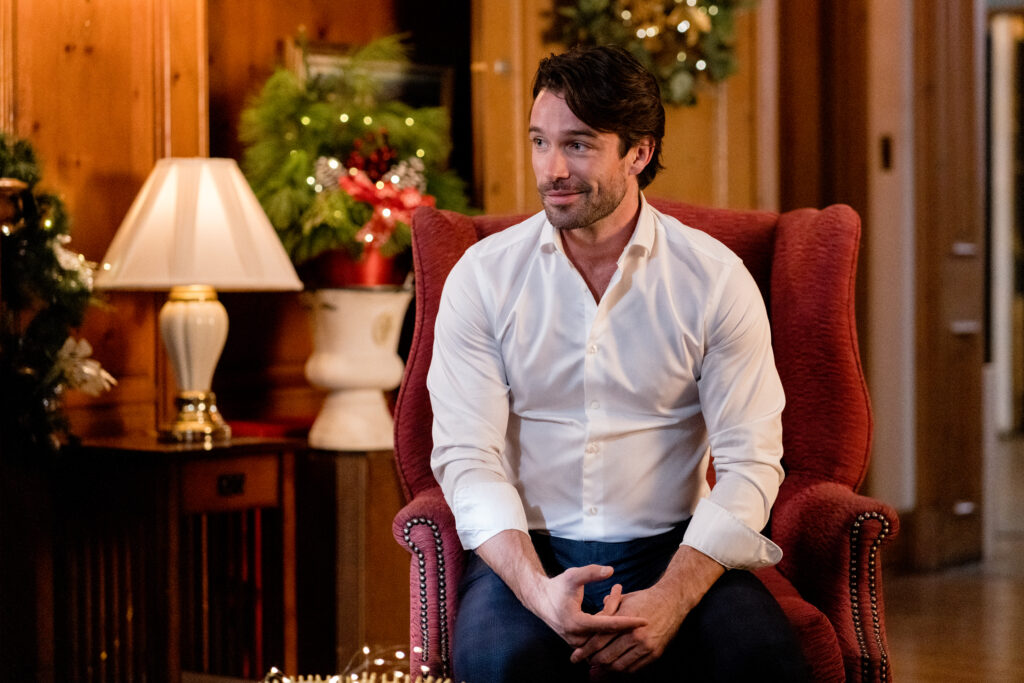 Joe Scarpellino sit in a library with a white shirt, smiling in a scene from the Hallmark movie Christmas with the Knightlys