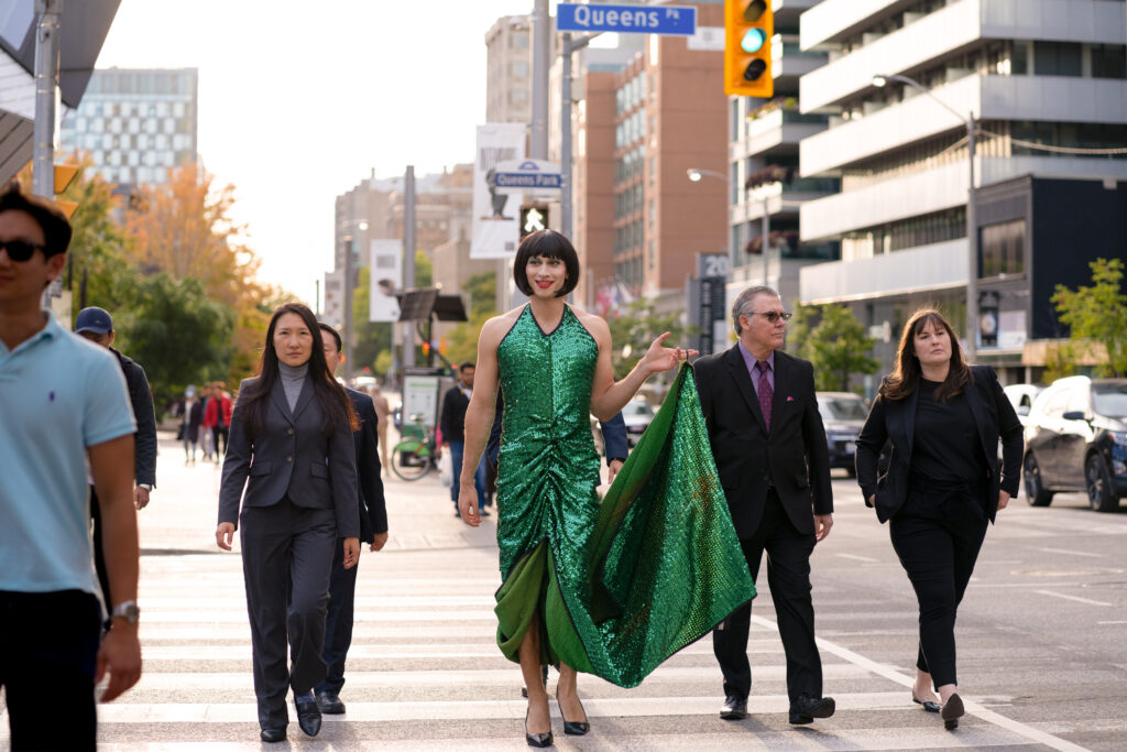 Ryan Ali walks down the streets of toronto in full Queen Tut drag, with a long green sequin dress and heels in a scene from the movie Queen Tut