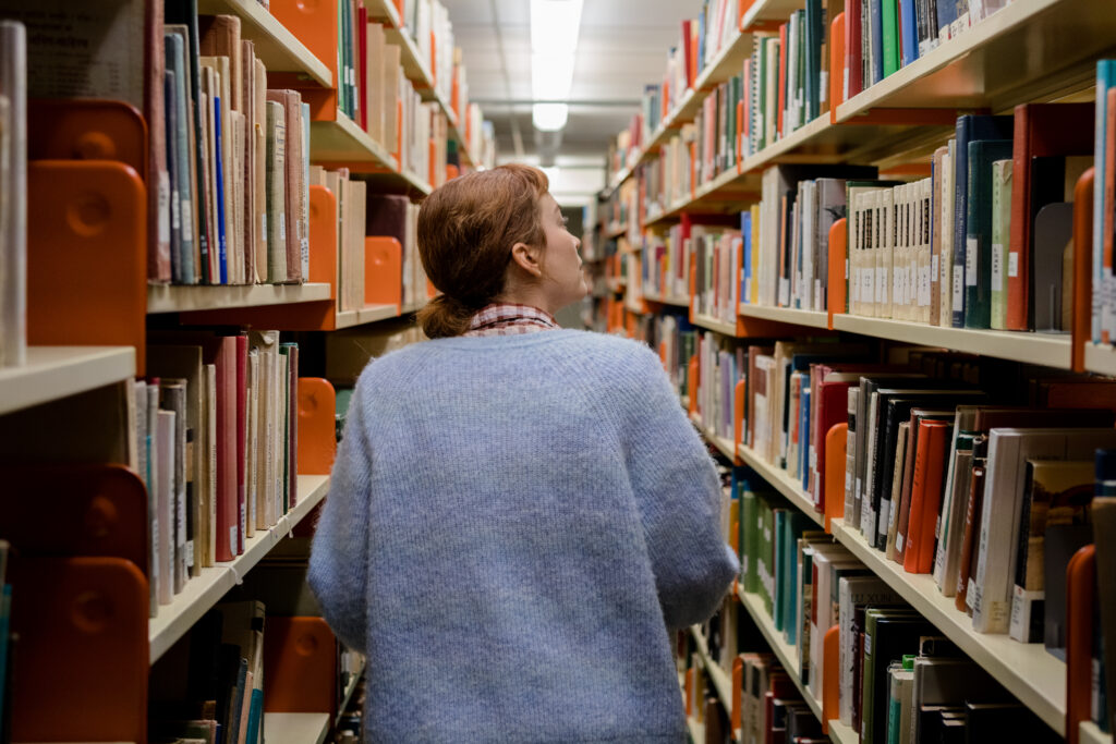 Britt Lower as Miriam walks through the library looking at books