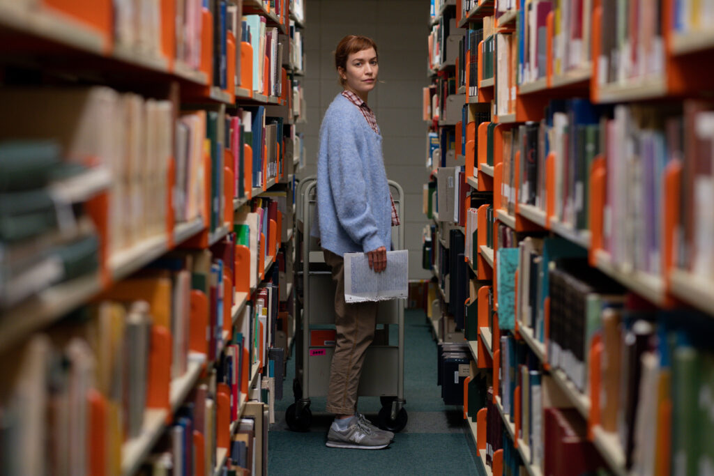 Britt Lower as Miriam stands in an aisle of books in a library looking to camera from a distance